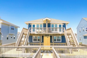 Een twee verdiepingen tellend strandhuis met een houten terras en een trap naar een balkon.
