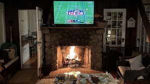 A cozy living room with a stone fireplace, a television displaying a sports game, and a comfortable seating area. - Adorable 3 bed cottage - 2 blocks to private beach; pond; AC, pets ok (Plymouth)