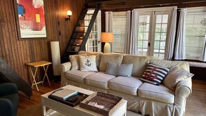A cozy living room with a beige sofa, a white coffee table, and a wooden wall with a framed abstract painting. - Adorable 3 bed cottage - 2 blocks to private beach; pond; AC, pets ok (Plymouth)