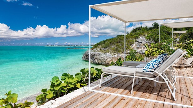 A wooden deck with lounge chairs and a canopy overlooking turquoise waters and a rocky coastline.