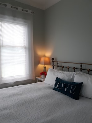 A neatly made bed with white linens and a pillow with the word "LOVE" on it, a bedside lamp, and a window with blinds.