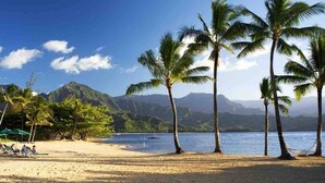 A beach with palm trees, a clear sky, and mountains in the background. - Stunning Views of Hanalei Bay and Bali Hai | Kauai North Shore (Princeville)
