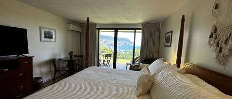 A hotel room with a large bed, a television, a desk with a chair, and a view of the sea through the window.