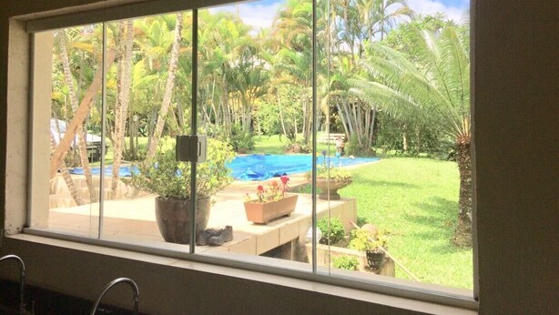 A window with a view of a backyard featuring a swimming pool, potted plants, and palm trees. - Mogi das Cruzes Farm - SP. - 90% Atlantic Forest - Cozy!!! (15 people) (Mogi das Cruzes)