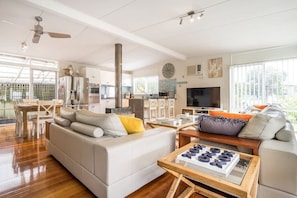 Ein modernes Wohnzimmer mit einer Couch, einem Couchtisch und einem Fernseher. Es gibt einen Essbereich mit Stühlen und einem Tisch. Der Raum verfügt über große Fenster und einen Deckenventilator.