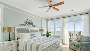 A bedroom with a bed, bedside tables, a ceiling fan, and a view of the ocean through large windows. - The Original Lulla-by the Sea Amazing Oceanfront Beach House W/Pool and Elevator (Carolina Beach)