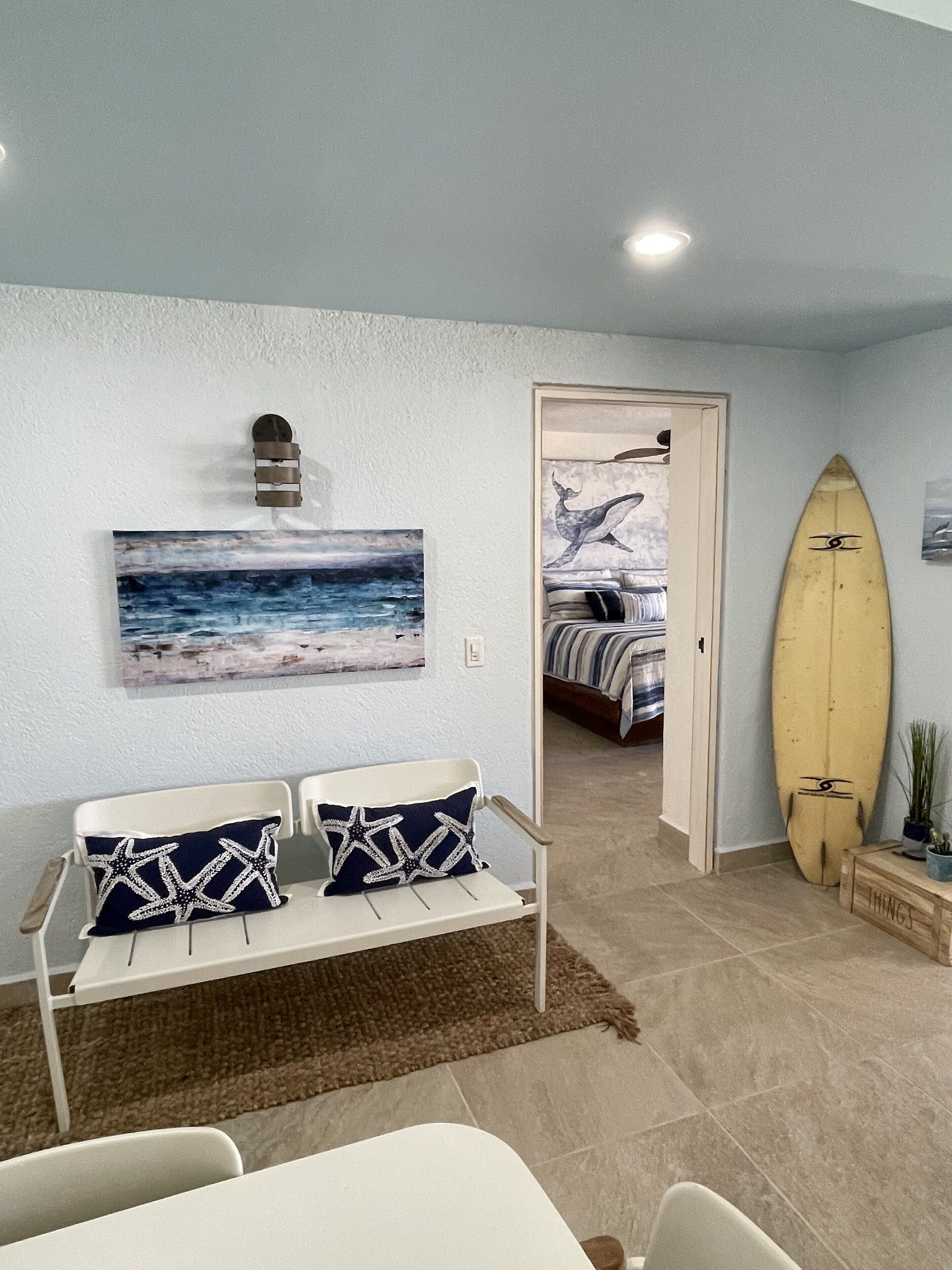 Une pièce avec un canapé blanc, une planche de surf appuyée contre le mur et un tableau représentant un paysage marin.