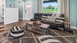 A living room with a sofa, ottoman, coffee table, and a view of a green landscape through a window. - 4th Nt Free! Waterfront Bungalow! Dock | Kayaks (Wills Point)