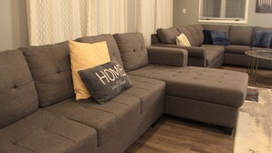 Un canapé modulable agrémenté d’un coussin orné de l’inscription « chez soi, quel bonheur », d’un pouf assorti et d’un tapis à motifs.