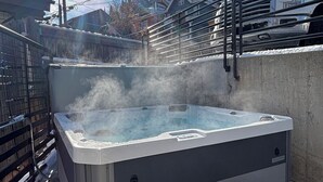 Un jacuzzi con vapor que se eleva, rodeado por un muro de concreto y baranda de metal, con cielo despejado y edificios al fondo.