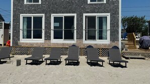 Un bâtiment à deux étages, à l’extérieur sombre et aux bordures blanches. Devant, on trouve six chaises longues alignées en rangée sur une zone sablonneuse.