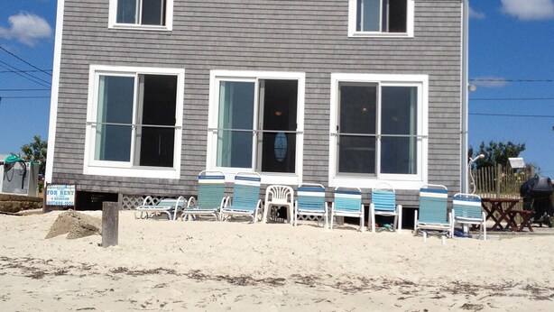 Une maison de plage à deux étages, dotée d’un balcon, d’une pancarte « À louer » et de chaises de plage devant.