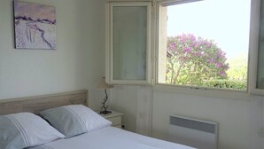 Een slaapkamer met een bed, een nachtkastje, een lamp, een raam met uitzicht op bomen en bloemen, en een schilderij aan de muur.