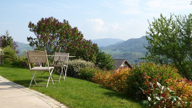 Een tuin met uitzicht op bergen, een klein huisje en twee stoelen op een verhard pad.