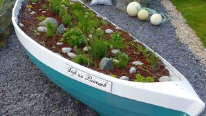 A boat-shaped planter with the inscription "Sigh na Boireach" filled with plants and rocks, placed on a gravel path with a large orange buoy nearby.
