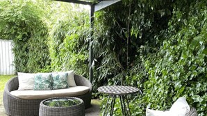 A covered outdoor seating area with wicker furniture, a small table, and a wall of greenery.