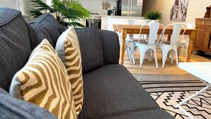 A modern living room with a grey sofa, patterned cushions, a dining area with white chairs, a wooden table, and a kitchen in the background.