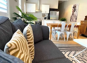 A modern living room with a grey sofa, patterned cushions, a dining area with white chairs, a wooden table, and a kitchen in the background.