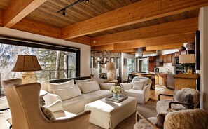 A spacious living area with a large sofa, armchairs, and a coffee table. There is a stone fireplace, a kitchen area, and large windows providing natural light. - Renovated Mountain-Modern Home, Hot Tub, Pass Views (Aspen)