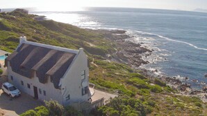 En kystejendom med et stort hus med udsigt over havet, en bil foran og frodig vegetation.