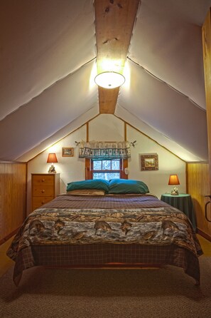 Een gezellig slaapkamer op zolder, met een schuine plafond, een bed met een beddeken met dessin, twee nachtlampjes, en een raam met uitzicht op buiten.