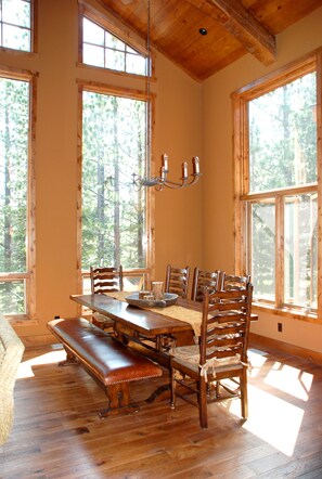 Enjoy meals in this spacious dining area with a long table and comfortable chairs, surrounded by large windows offering views of the forest.