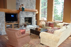 Relax in this inviting living room with a fireplace, comfortable seating, and a TV, plus a window with a view of the surrounding trees.