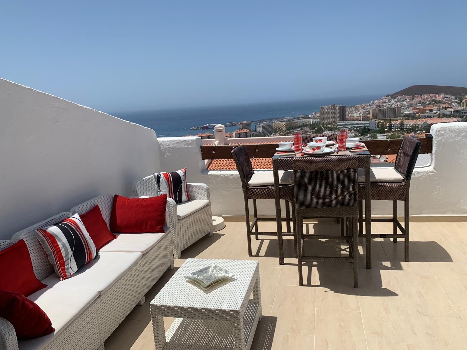 A terrace with a white sofa, dining table, and chairs, overlooking a coastal cityscape.