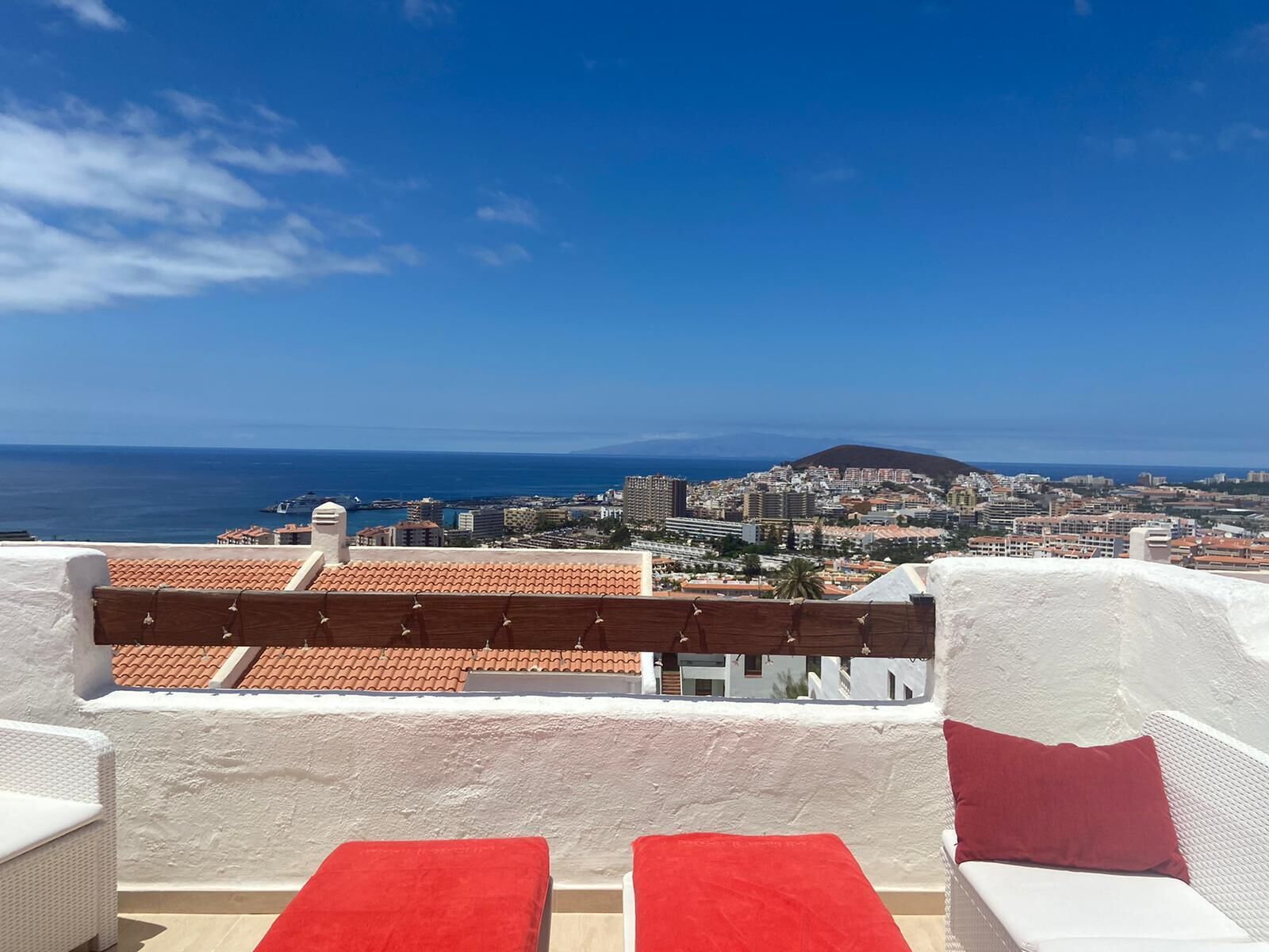 A terrace with a view of a coastal city, featuring a white wall and red cushions.