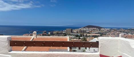 A terrace with a view of a coastal city, featuring a white wall and red cushions.