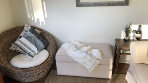 A cozy living room with a wicker chair, a cushioned bench, a small side table with a bottle and candle, and a framed picture on the wall. - Fairmont Cottage (Woodhill)