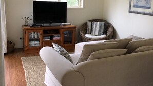 A living room with a beige sofa, a television on a wooden stand, a wicker chair, and a large window. - Fairmont Cottage (Woodhill)