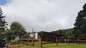 A grassy area with a wooden fence, a building with a flat roof, and trees in the background. - Fairmont Cottage (Woodhill)