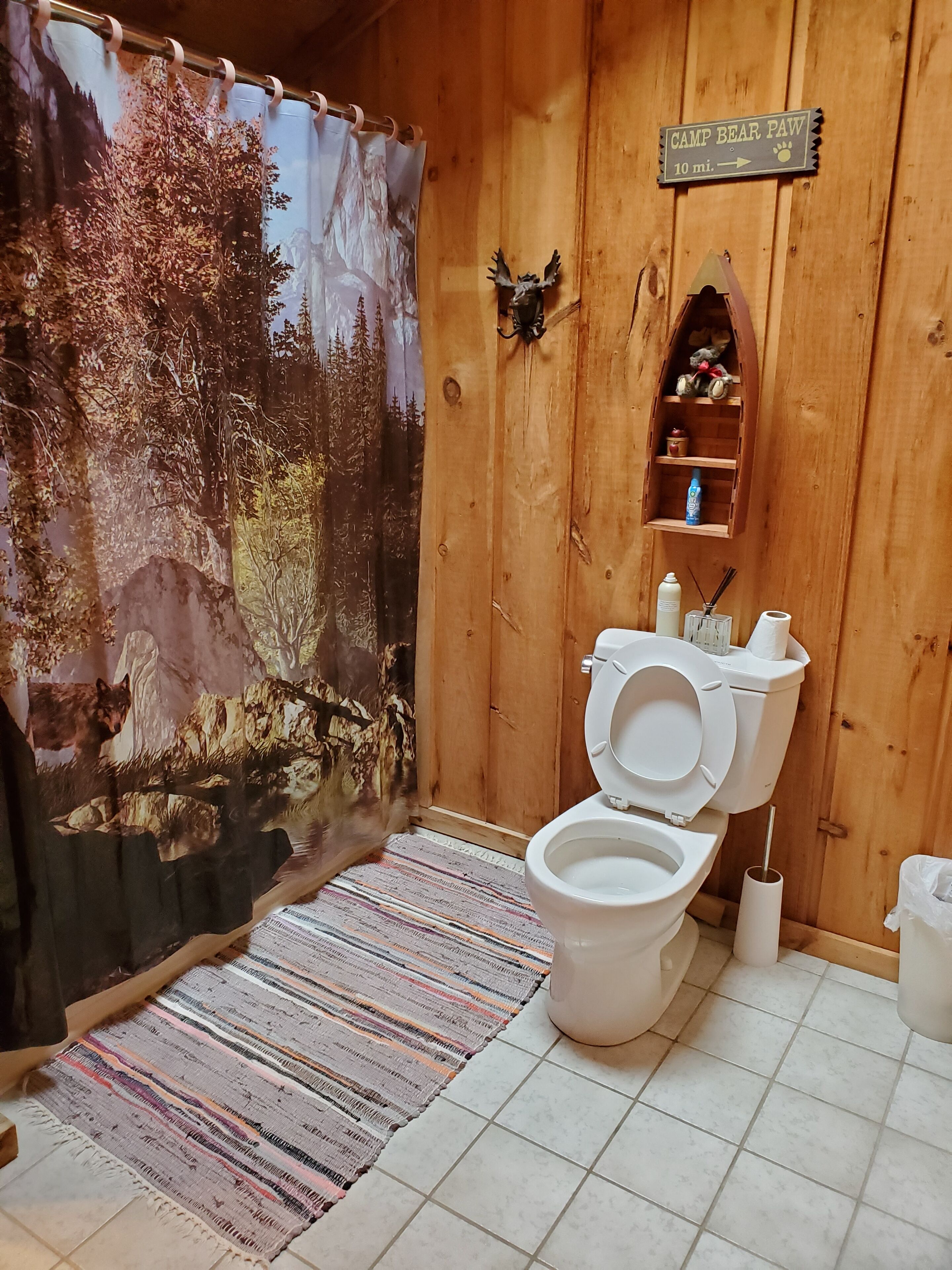 Ett badrum i lantlig stil med ett duschdraperi med skogsmotiv, en toalett och en liten hylla med toalettartiklar.