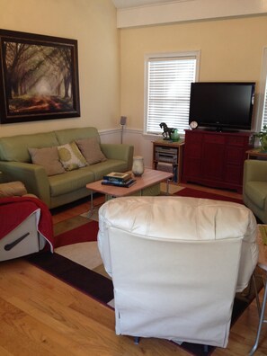 Relax in this inviting living room with a sofa, armchair, and TV, surrounded by artwork and natural light.