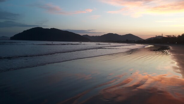 Una playa con aguas tranquilas y montañas lejanas bajo un cielo al atardecer.