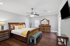 A bedroom with a wooden bed, a dresser, a bench, a ceiling fan, and a flat-screen TV mounted on the wall.