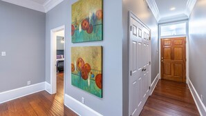 A hallway with hardwood floors, a white door, and two large abstract paintings on the wall. - Garden District Upscale Escape w/Gated Off-St Parking & Streetcar Access (New Orleans)