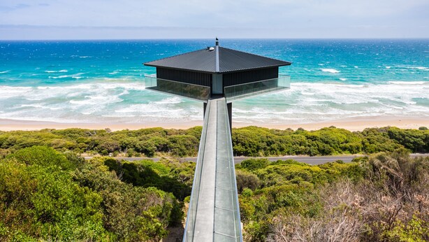 A modern building with a flat roof, situated on a high structure overlooking a beach and the ocean.