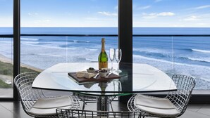 A table with a bottle, glasses, and food items, overlooking a beach and ocean view.