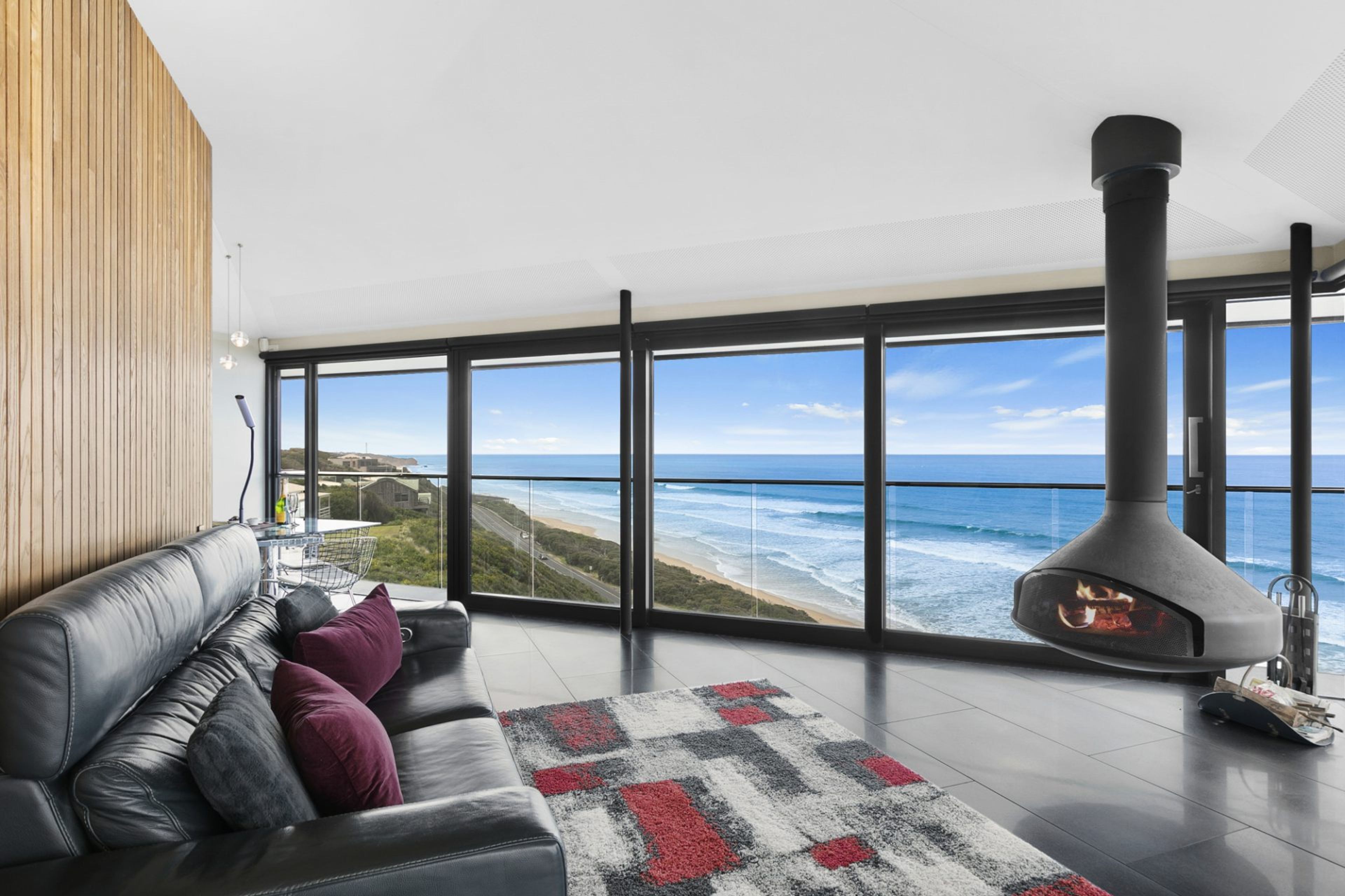 A modern living room with a leather sofa, a patterned rug, and a large window overlooking a beach and ocean.