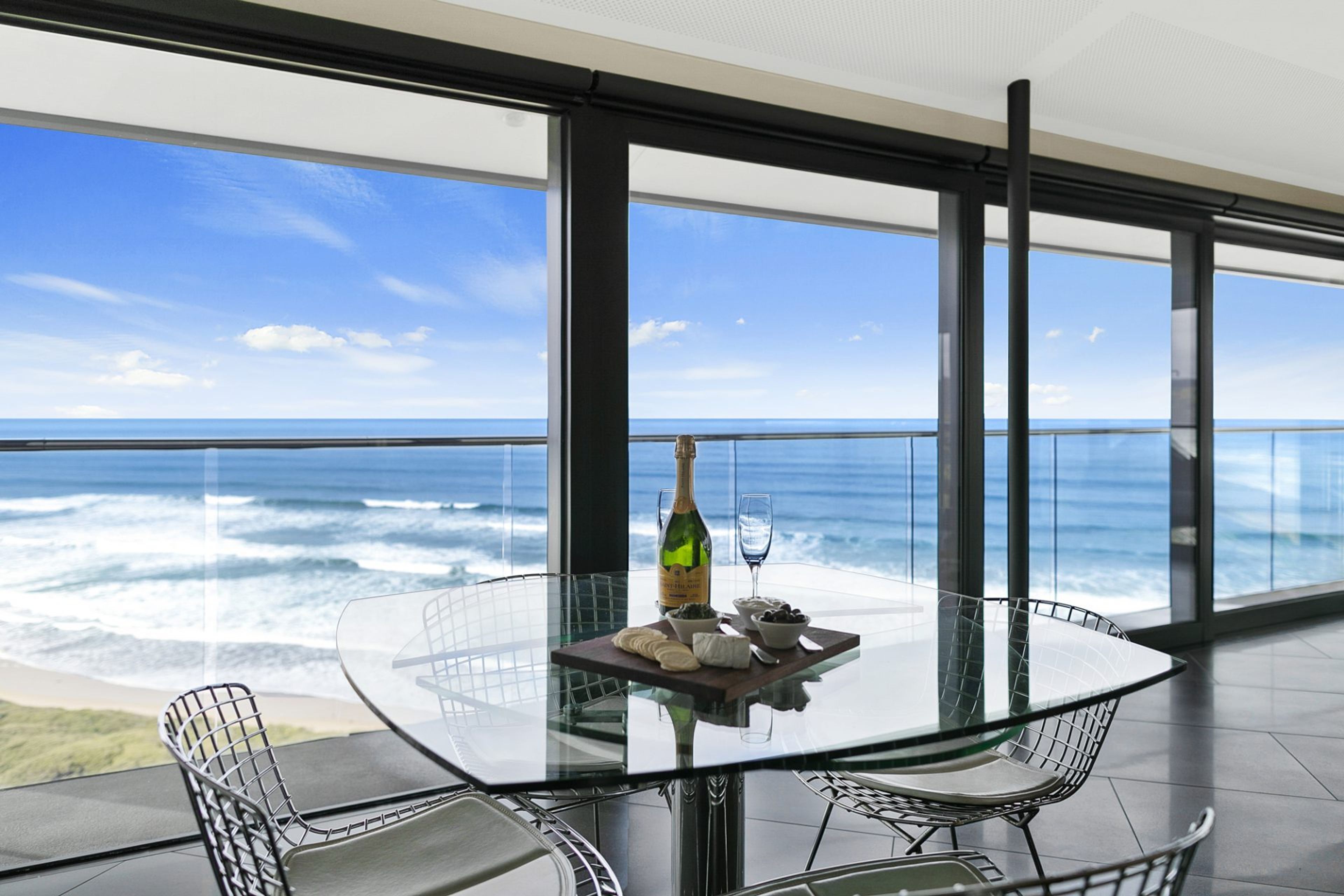 A glass table with a bottle and glass, surrounded by metal chairs, overlooking a view of the ocean.