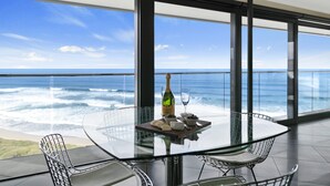 A glass table with a bottle and glass, surrounded by metal chairs, overlooking a view of the ocean.
