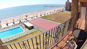 En balkon med udsigt over en strand, en tennisbane og en swimmingpool.