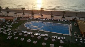 Et poolområde med liggestole og parasoller, en swimmingpool og havudsigt i solnedgang.