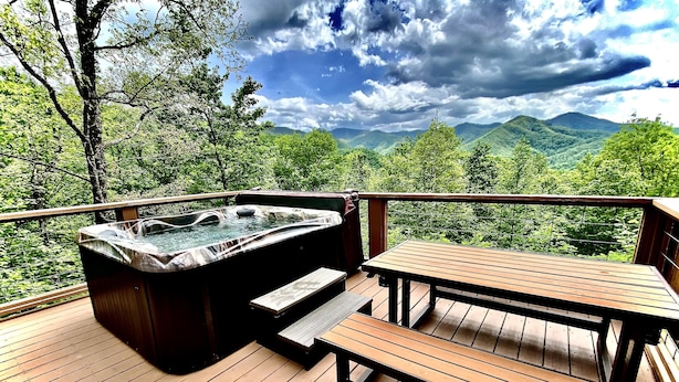Un jacuzzi en una terraza de madera con una mesa y un banco, rodeado de vegetación exuberante y montañas al fondo.