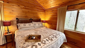 Un dormitorio de cabaña de madera con una cama, dos lámparas de noche, una mesita de noche de madera y un ventanal con cortinas.