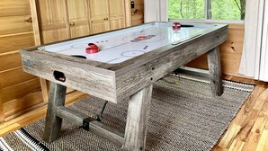 A wooden-finished shuffleboard table with a glass top, set in a room with wooden walls and a large window overlooking greenery.