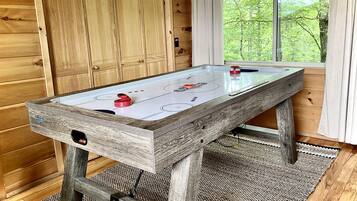 Puisella viimeistelyllä varustettu shuffleboard-pöytä, jossa on lasipintainen pelialusta. Pöytä on sijoitettu huoneeseen, jossa on puuseinät ja suuri ikkuna, josta avautuu näkymä vehreään maisemaan.