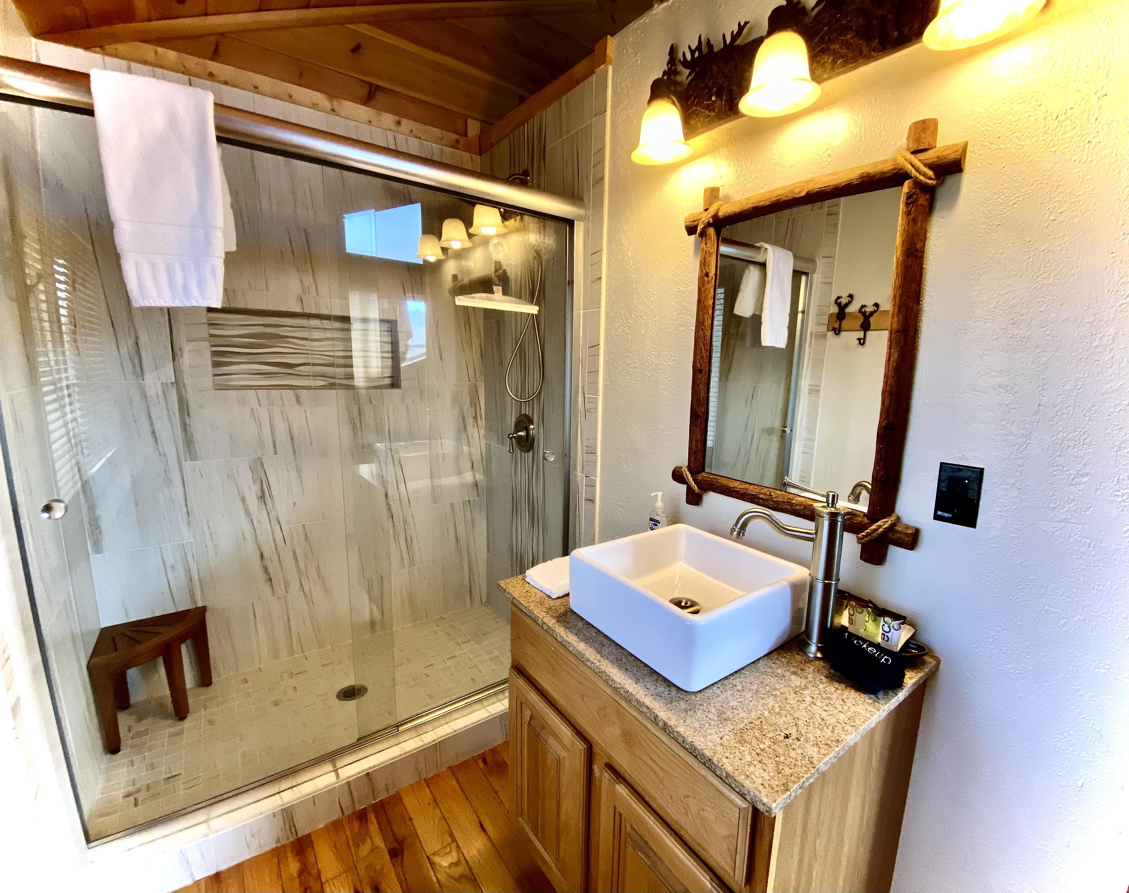Un baño con ducha envuelta en vidrio, un lavabo blanco, armarios de madera y un espejo con marco de madera.